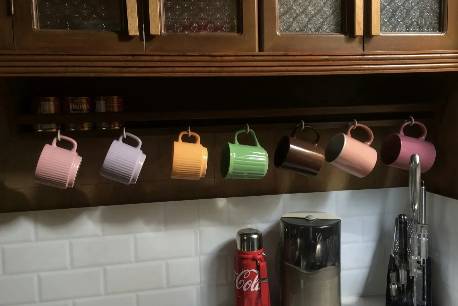Pastel mugs hanging in a row from hooks under dark wood upper cabinets above a white tile backsplash