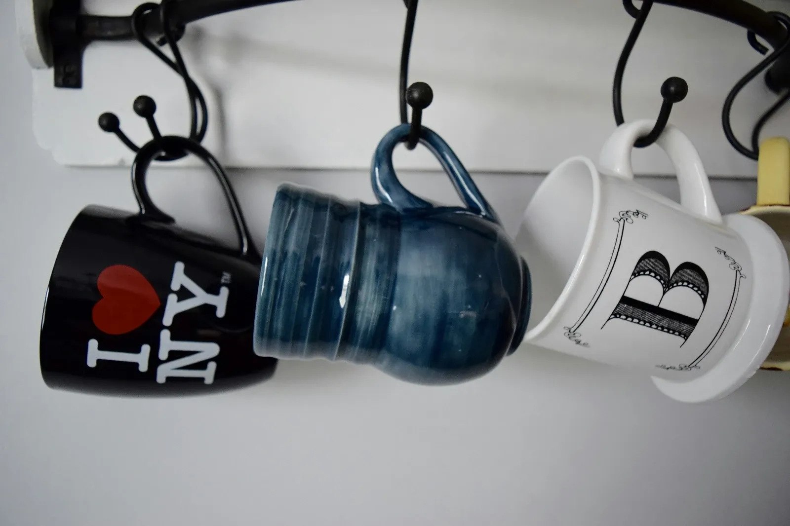 Three ceramic mugs in black, navy blue and white hanging from black hooks under a white shelf