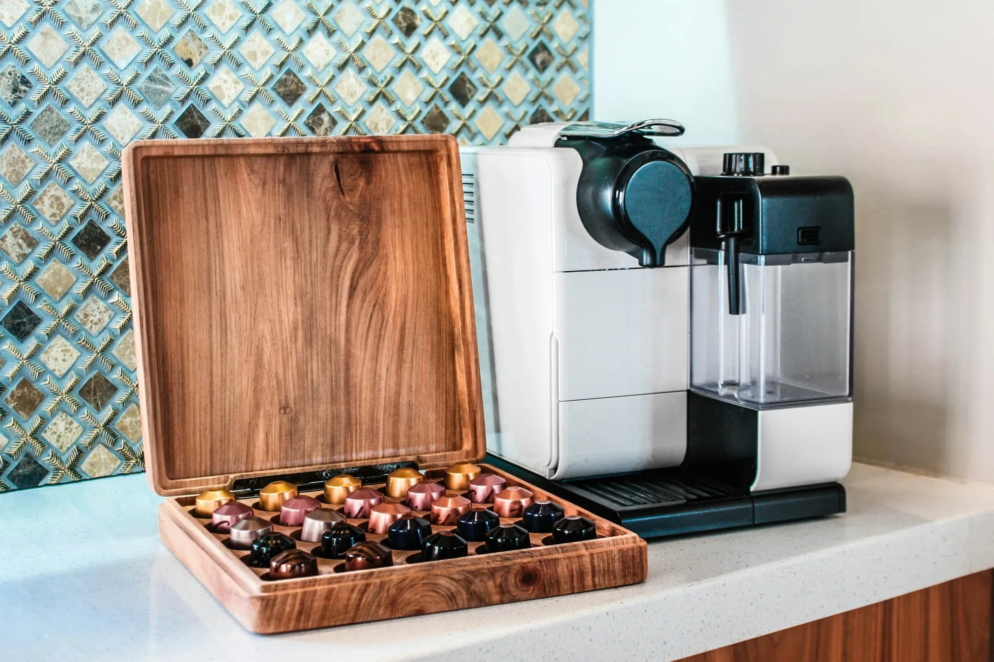White Nespresso capsule machine with a milk carafe beside an open wooden capsule box on a white counter with a mosaic tile backsplash