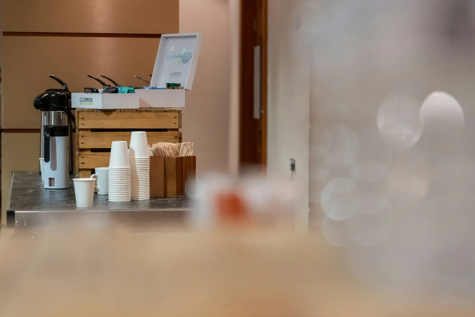 Office coffee cart with a pump airpot, stacks of paper cups and a wooden crate of supplies by a doorway