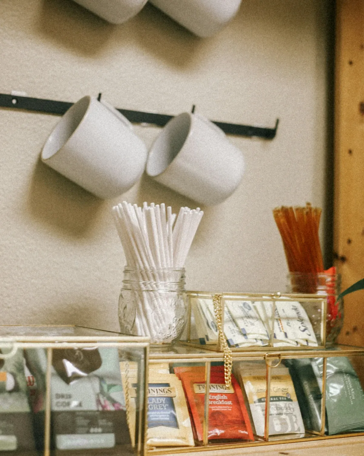 White mugs on black wall hooks above a jar of straws and stirrers and brass-framed boxes of tea bags on a wood counter