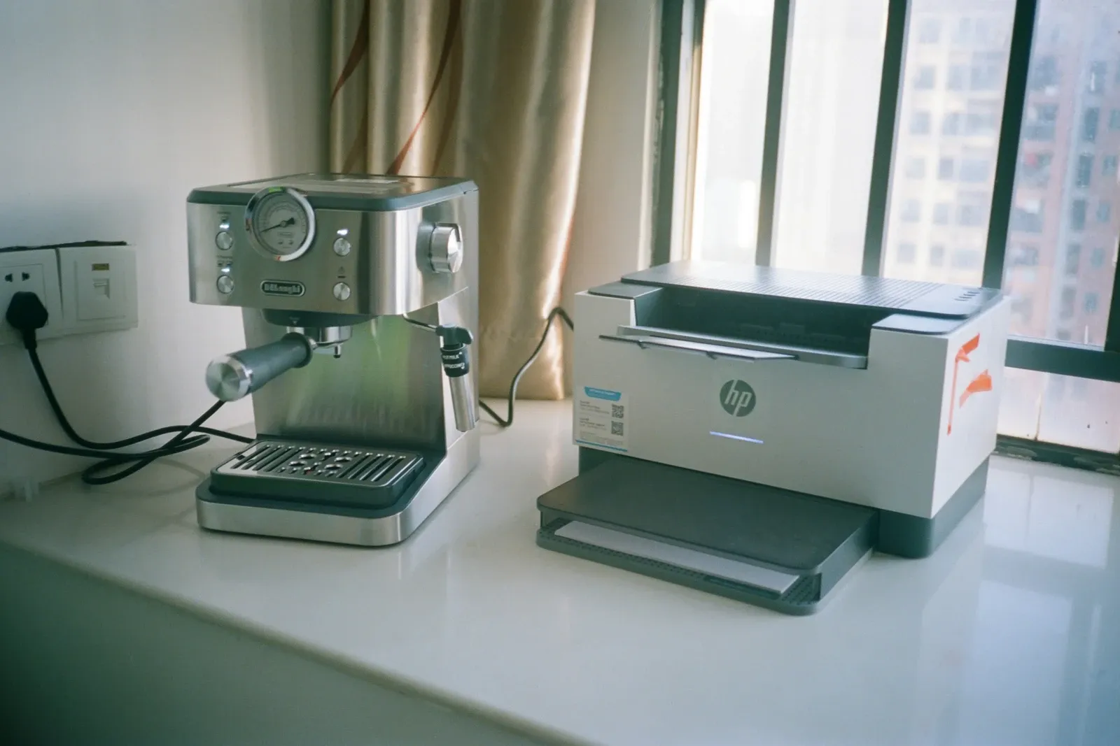 Stainless espresso machine on a white windowsill counter next to a compact laser printer in a high-rise office