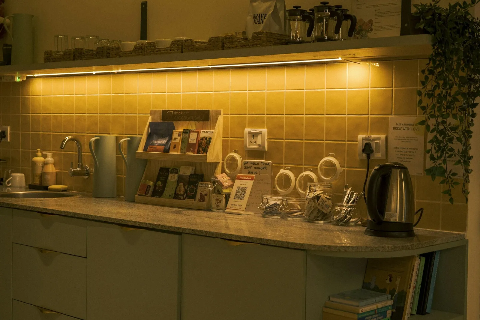 Kitchen counter corner with tea and coffee supplies, a kettle and warm under-cabinet lights on a tiled wall