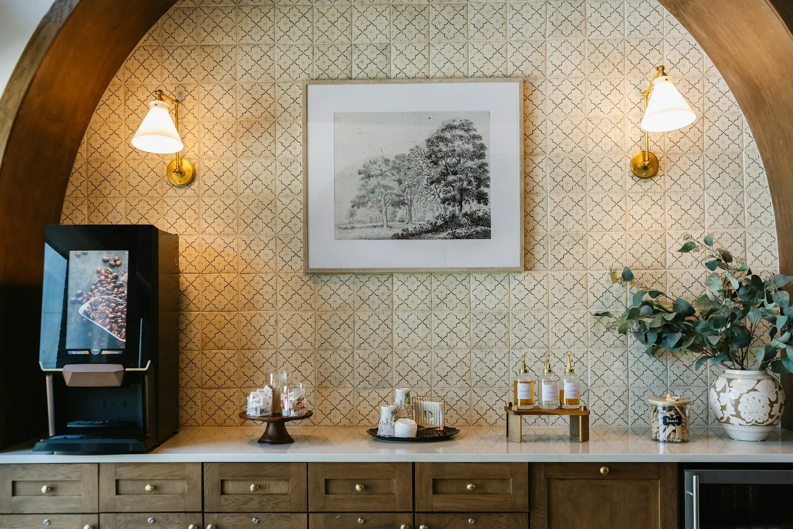 Office lounge coffee station with a bean-to-cup machine, framed print and wall sconces in an arched tile niche over wood drawers