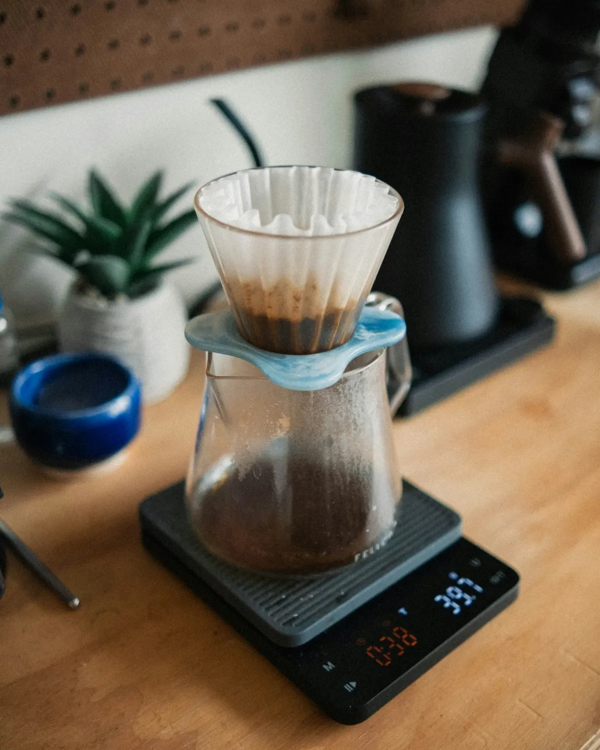 Pour-over dripper and glass server on a digital coffee scale on a wooden counter with a plant behind