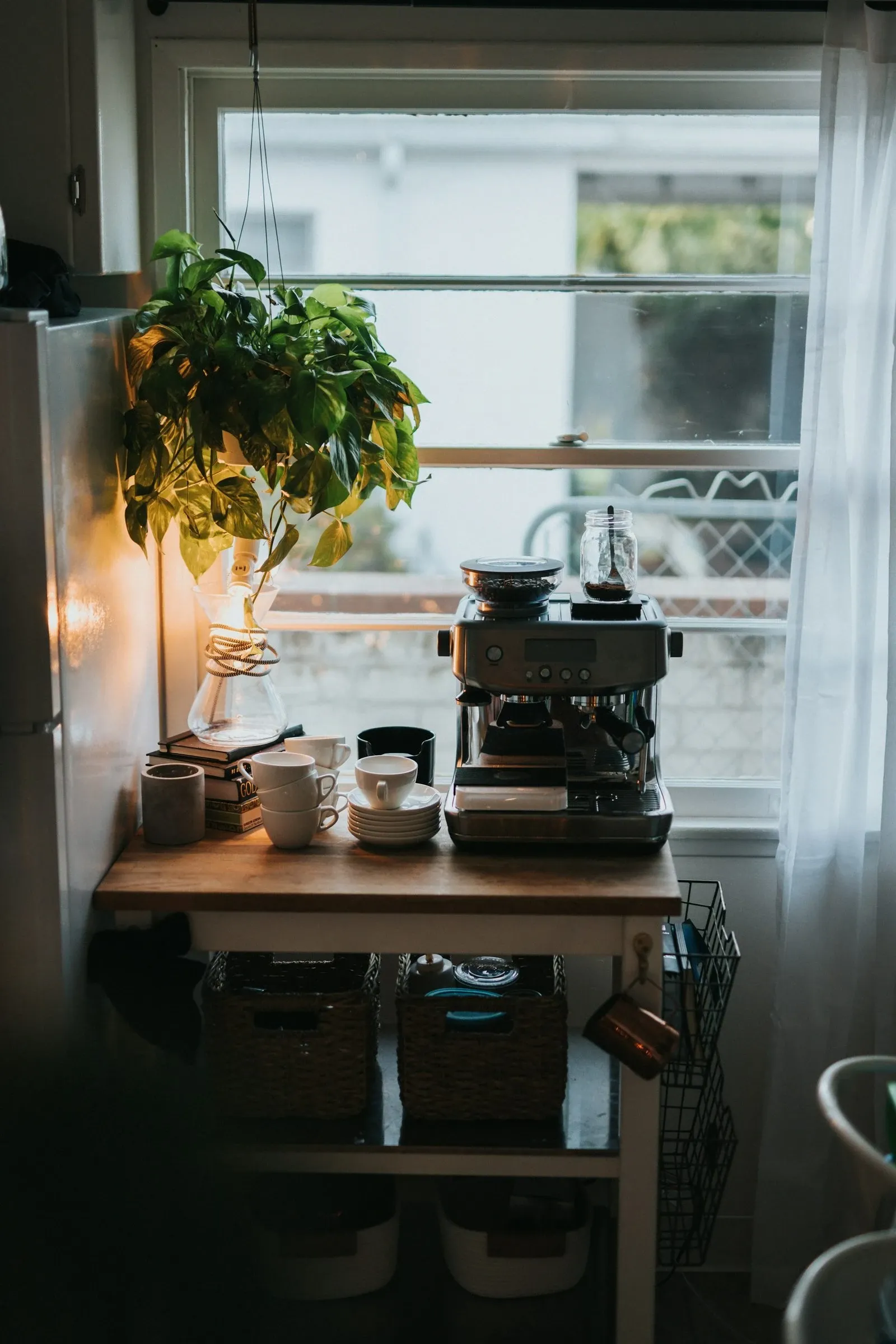 Wooden rolling cart by a window holding an espresso machine, cups and a trailing plant