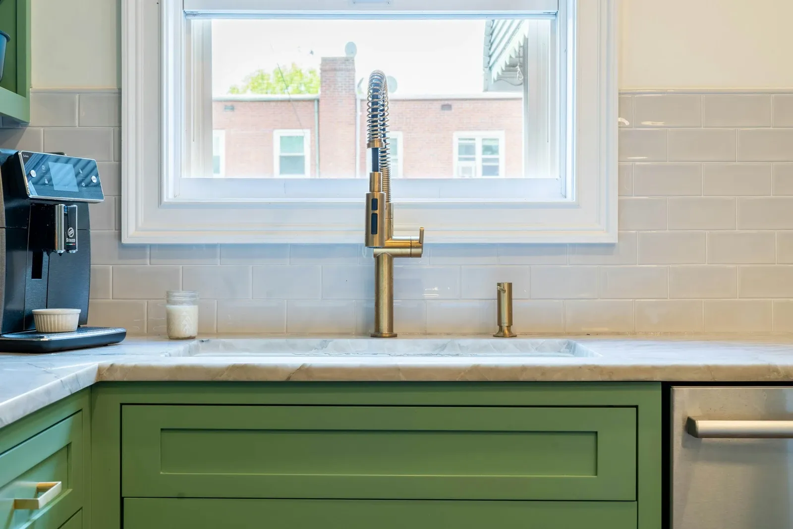Sage green cabinets under a white counter with a gold faucet and a window above the sink
