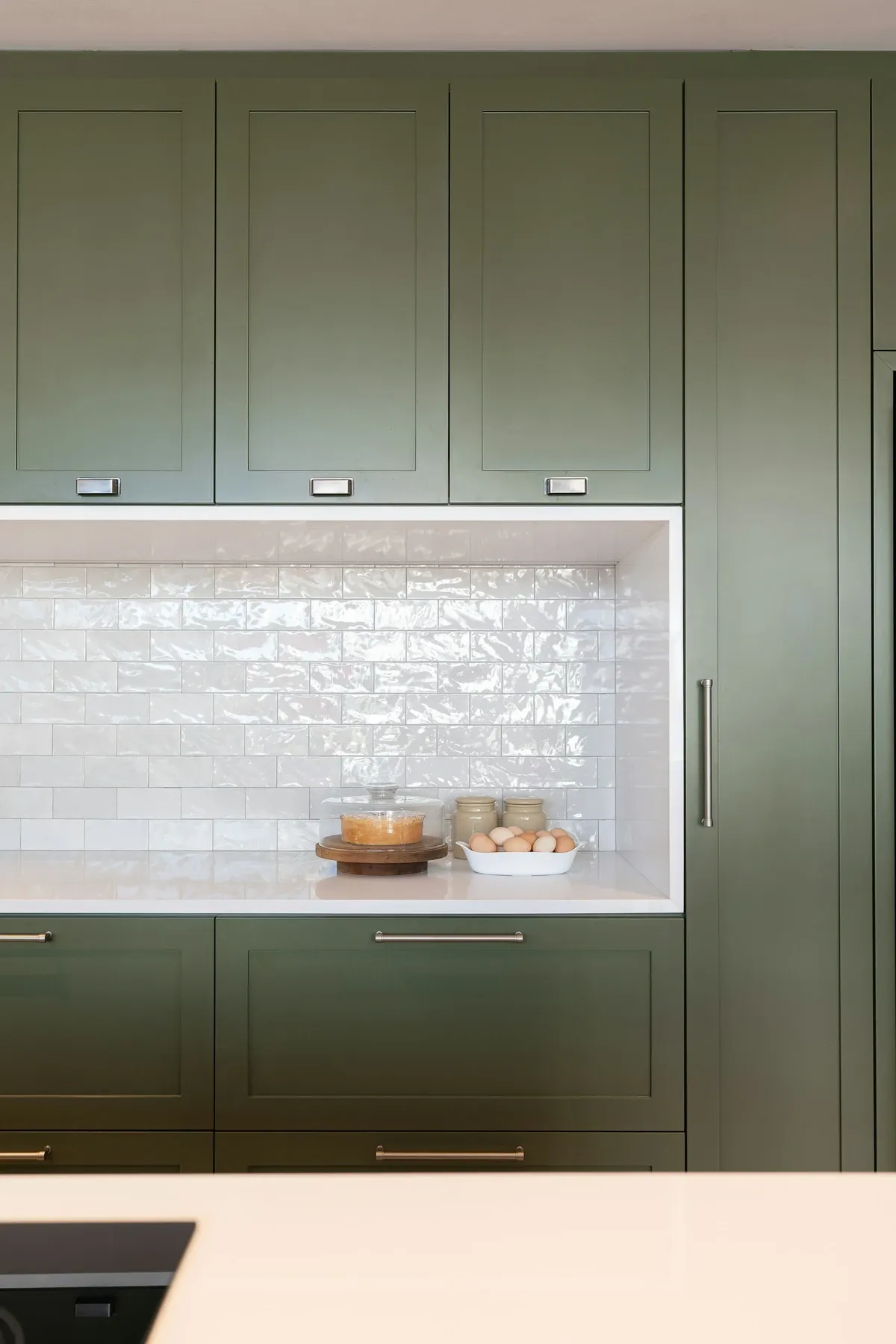 Sage green floor-to-ceiling cabinets around a built-in niche lined with white glossy tile, holding a small tray and a cup