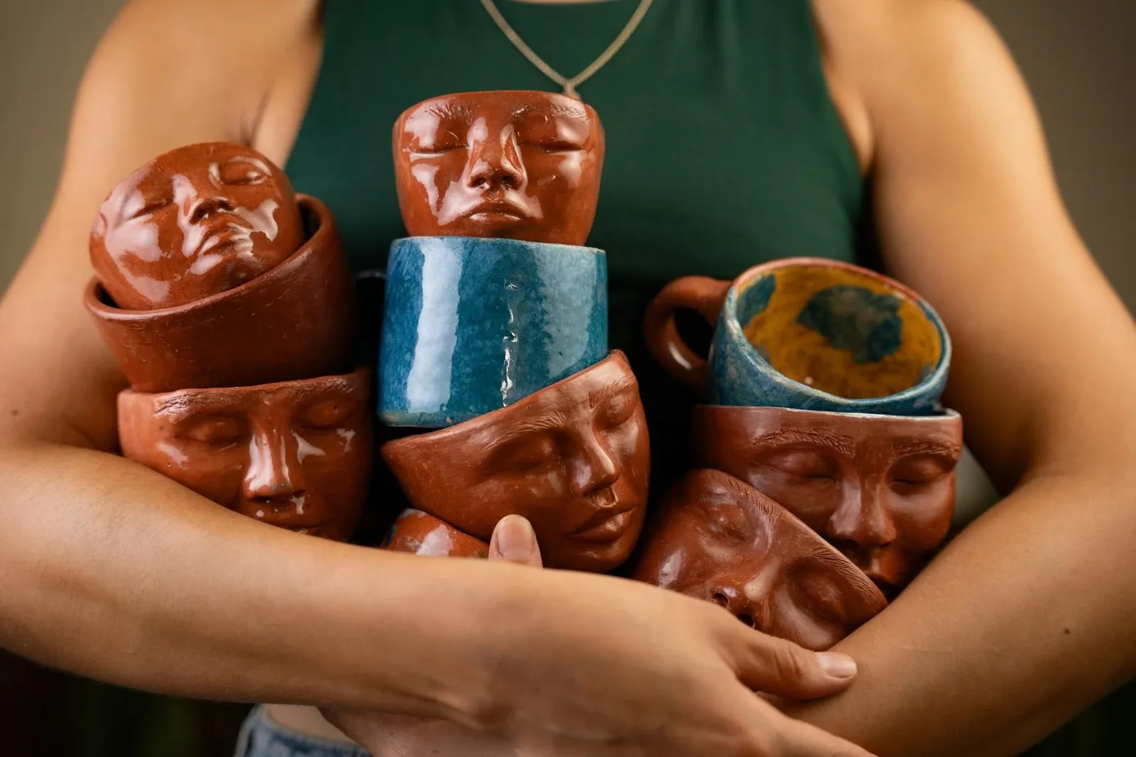 Person holding an armful of handmade ceramic cups sculpted with faces in red, blue and brown glazes