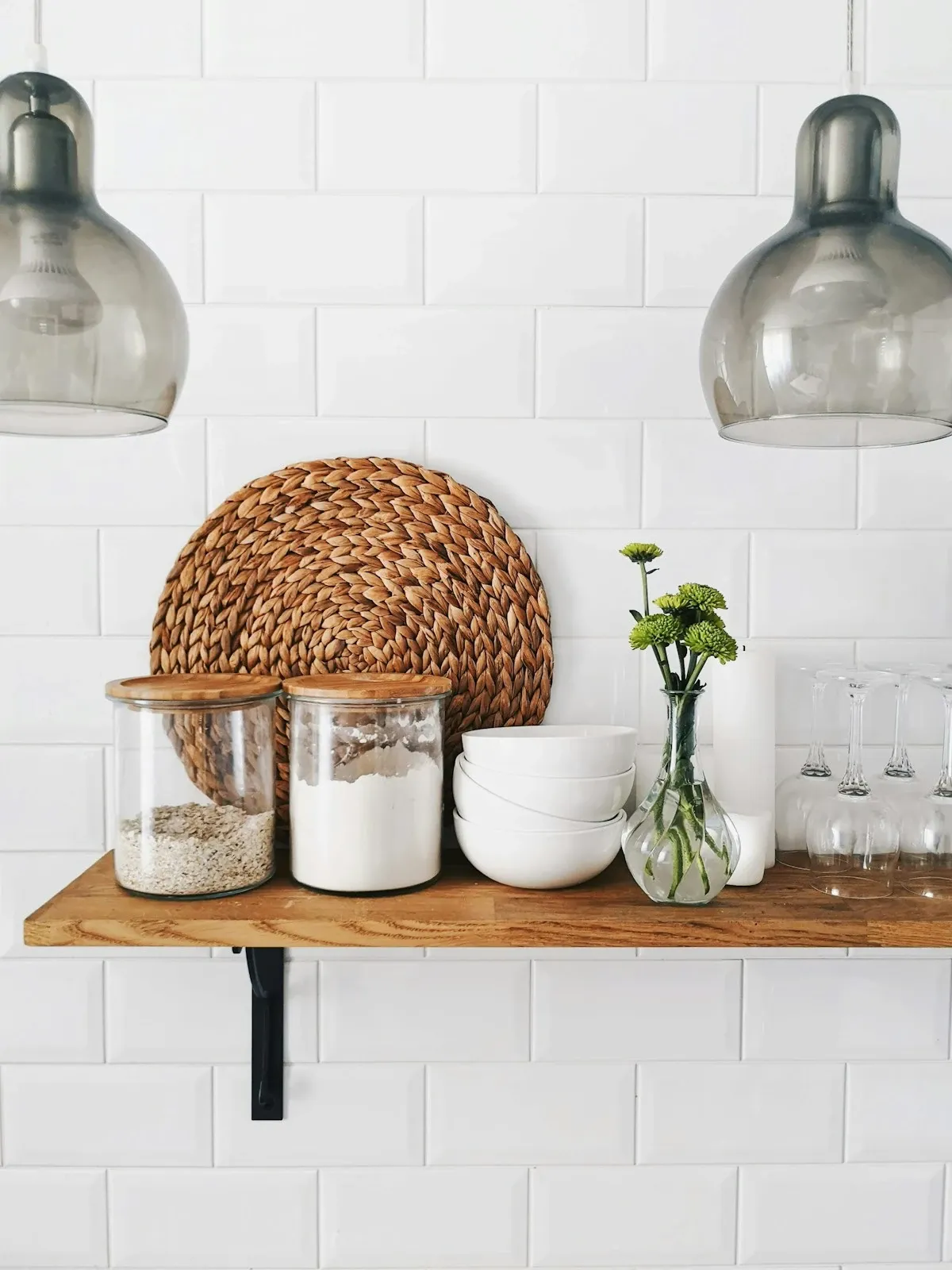 Single wood floating shelf with glass jars and a woven trivet on white subway tile, with pendant lights above