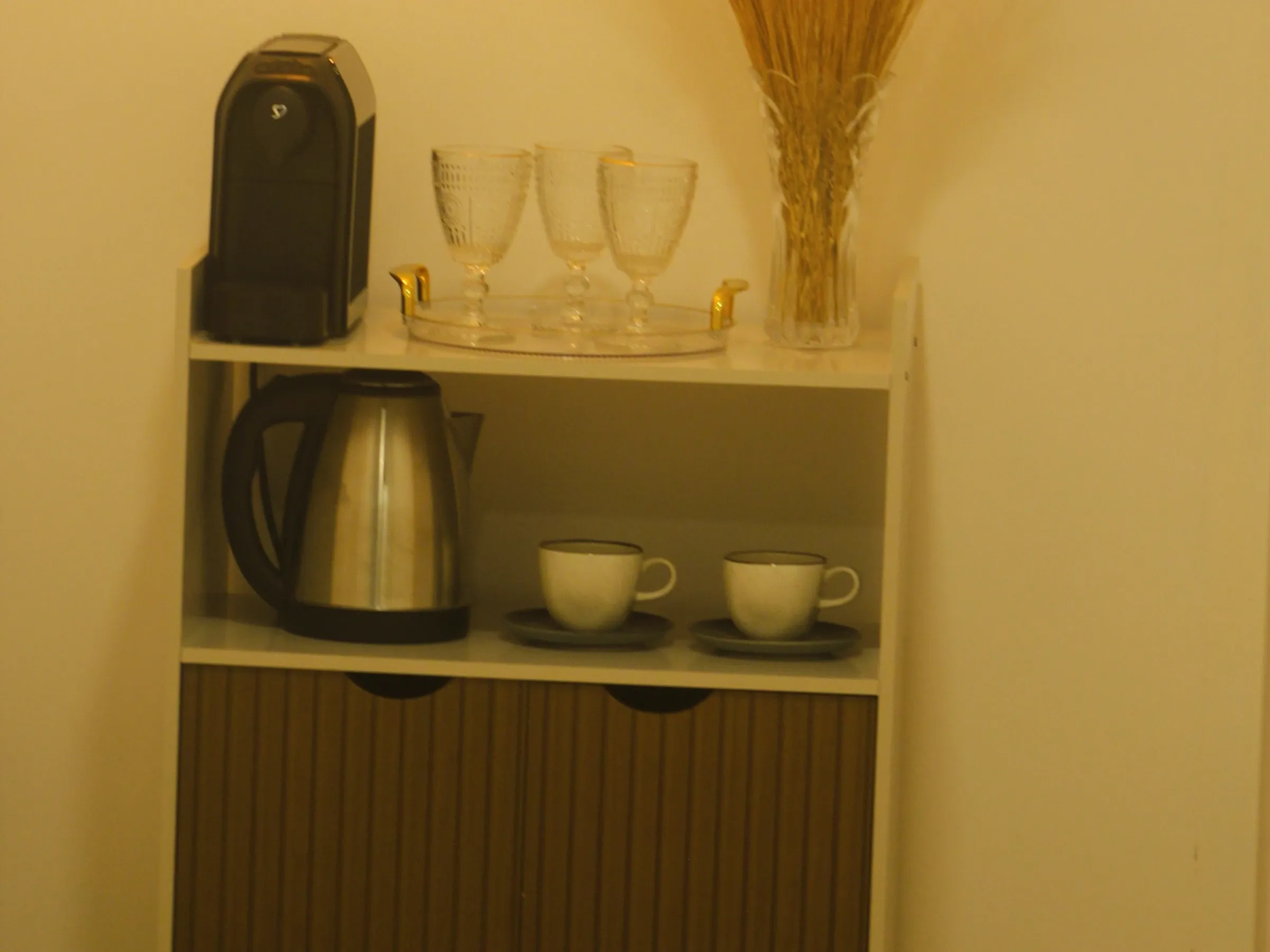 Small white shelf cabinet with a pod coffee machine, a gold tray of glasses, a steel kettle and two cups on saucers against a warm wall