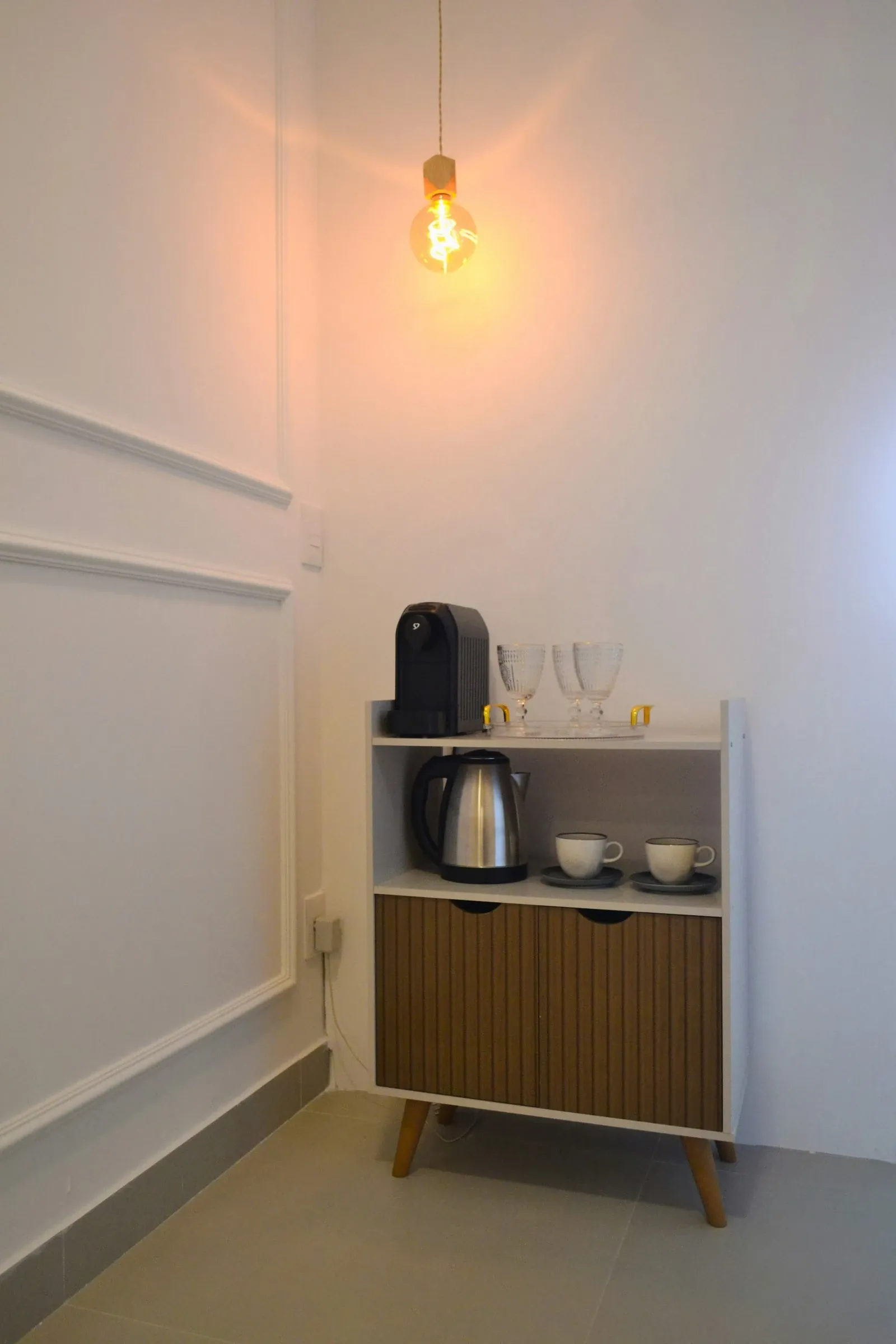 Cream two-tier cabinet holding a kettle and coffee maker on top, cups and glasses on the open shelf