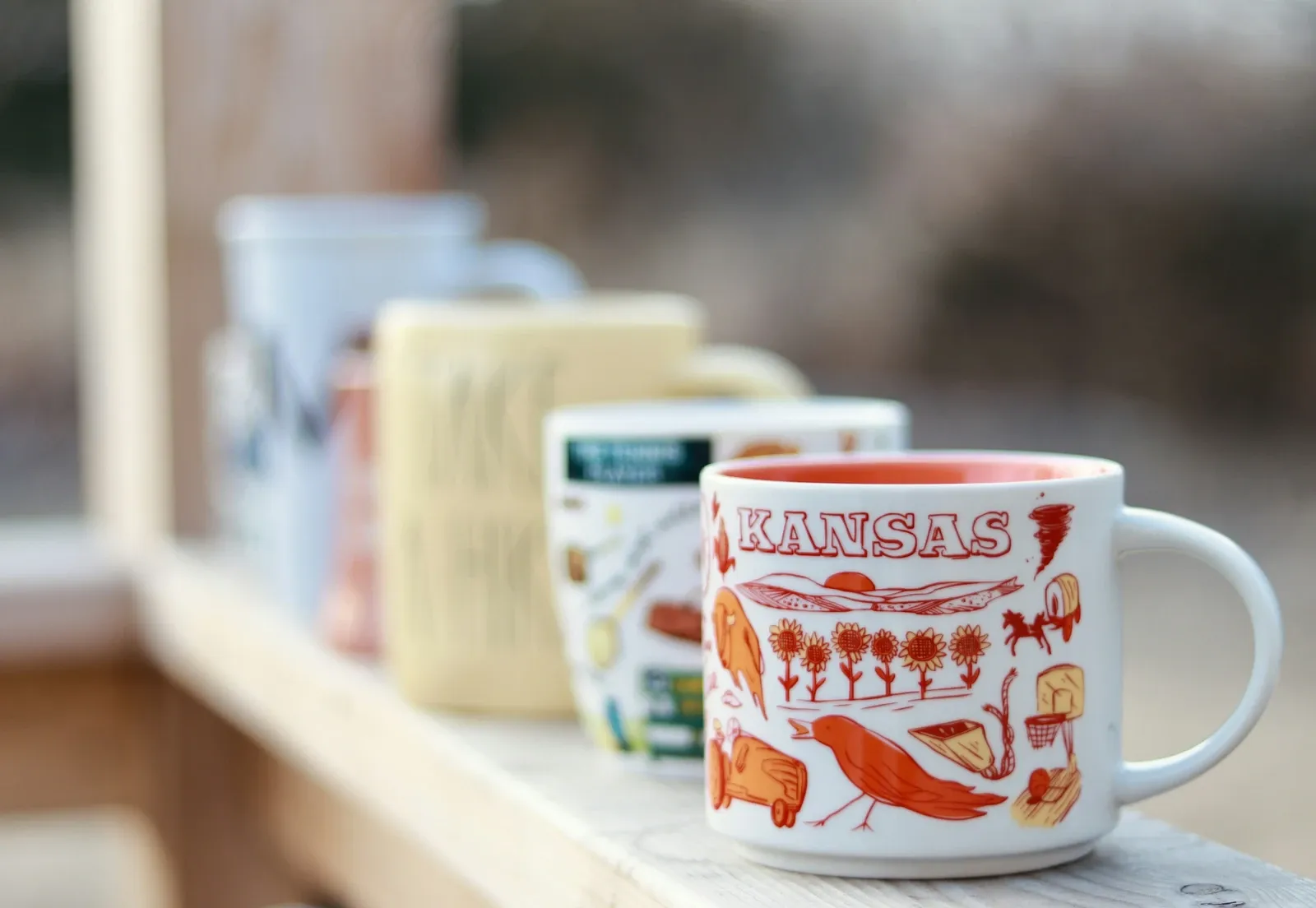 Row of souvenir city mugs with colorful illustrations lined up on a light wood windowsill shelf