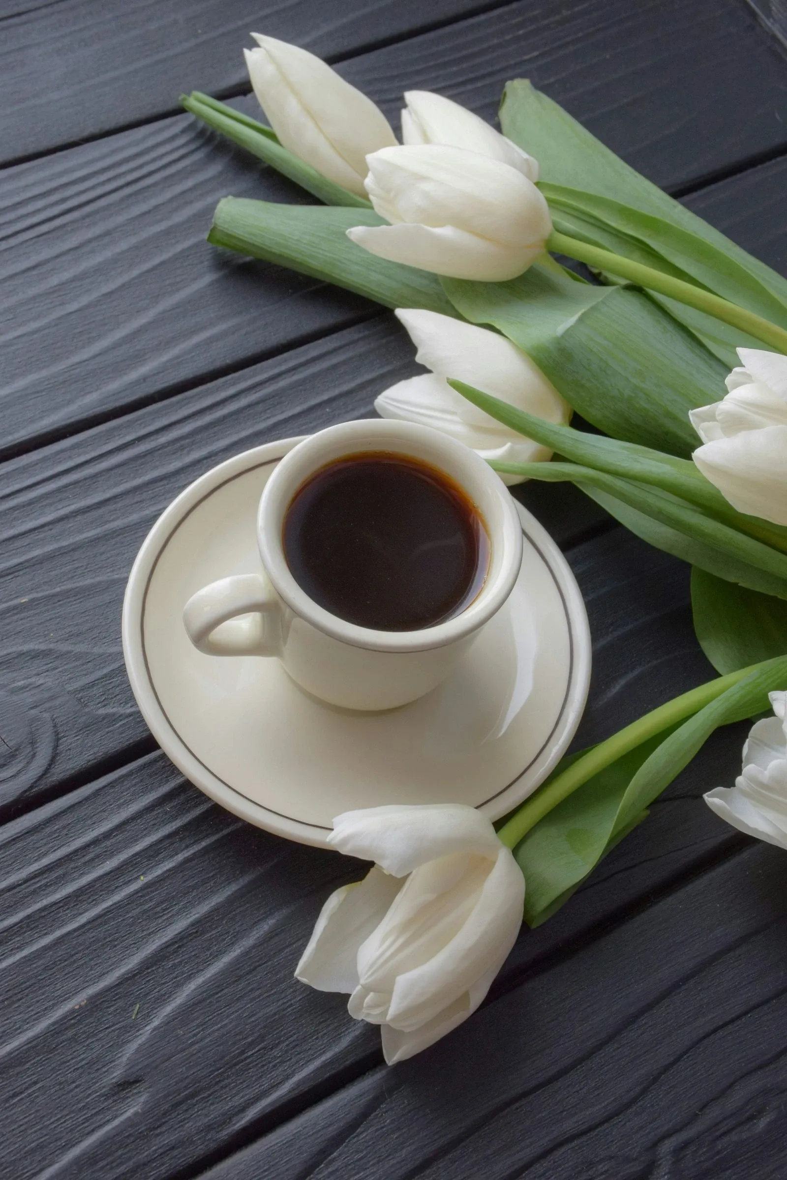 Espresso cup on a white saucer beside a bunch of white tulips on dark wood