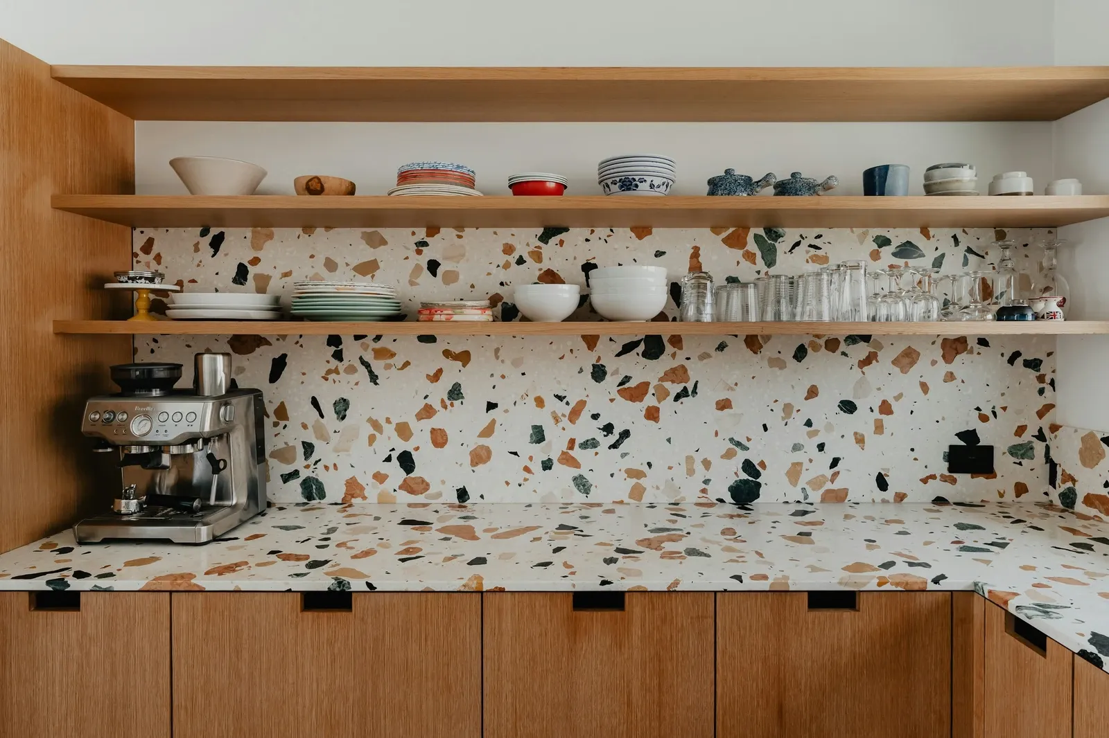 Espresso machine on a terrazzo counter under light wood open shelves stacked with bowls, cups and glasses