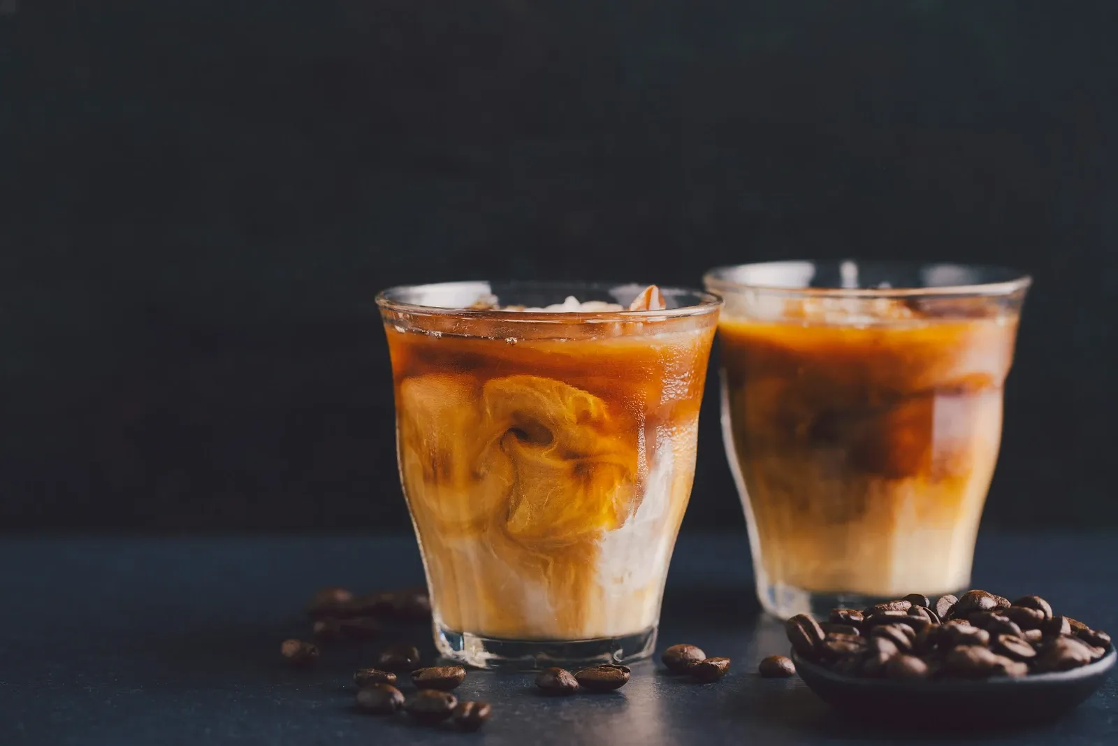 Two glasses of iced coffee with milk swirling through on a dark table with coffee beans