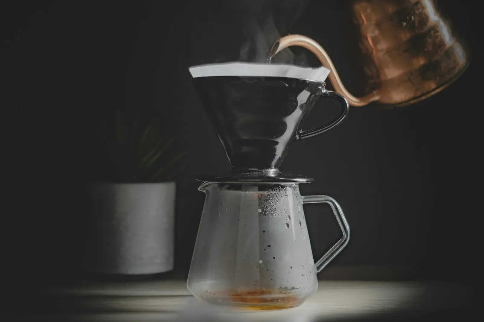 Copper gooseneck kettle pouring into a black V60 dripper on a glass server on a dark counter