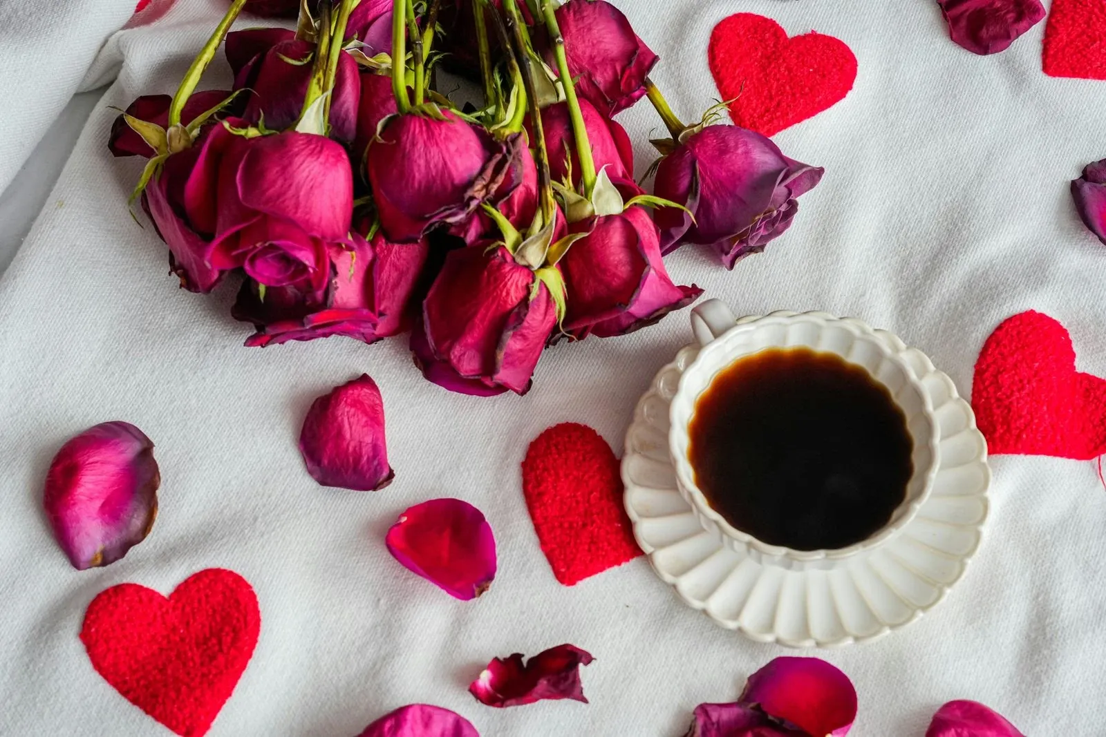 Coffee cup on a white cloth surrounded by dried pink roses and red paper hearts