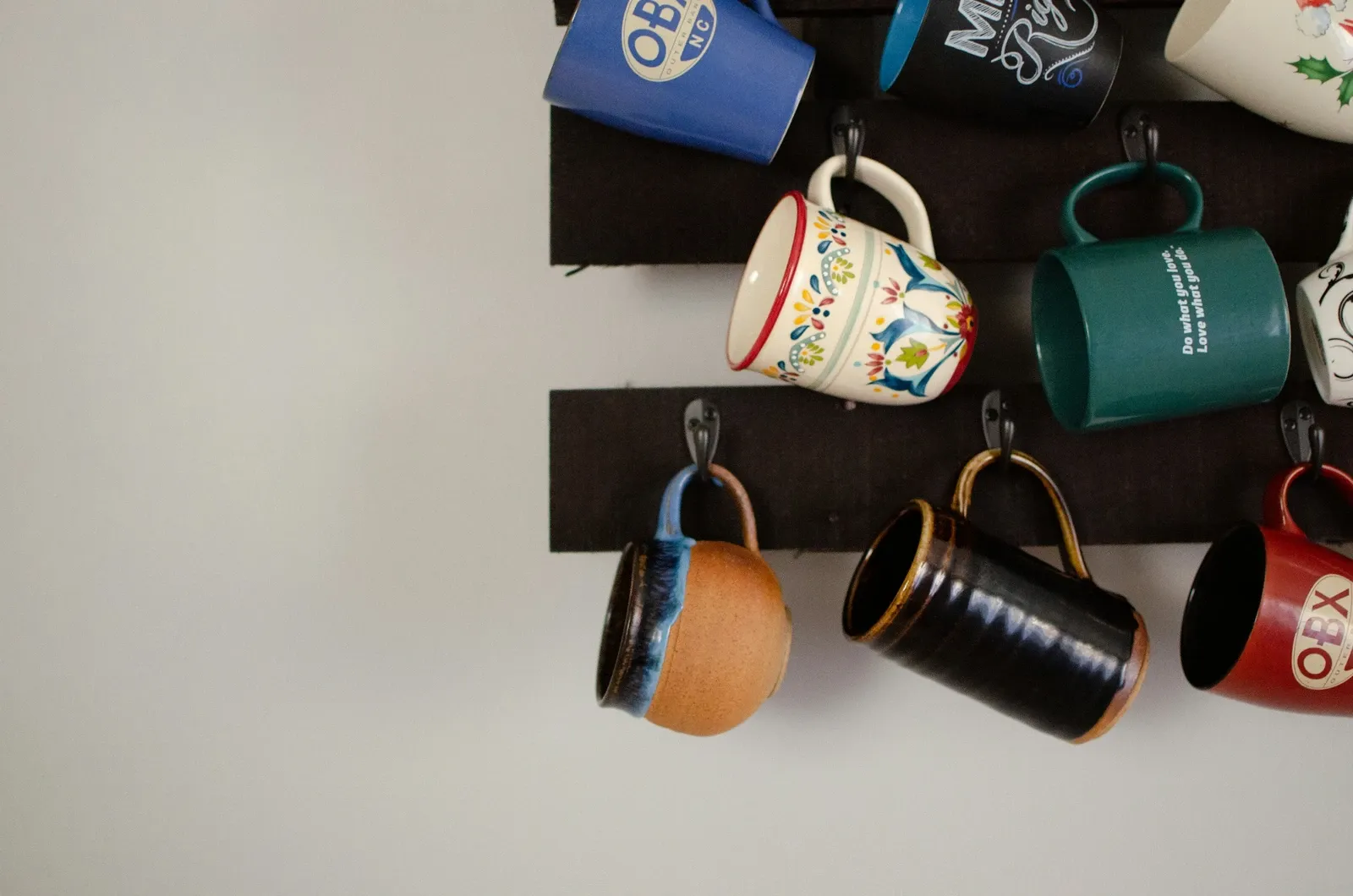 Colorful ceramic mugs hanging on black hooks from dark wood boards mounted on a white wall