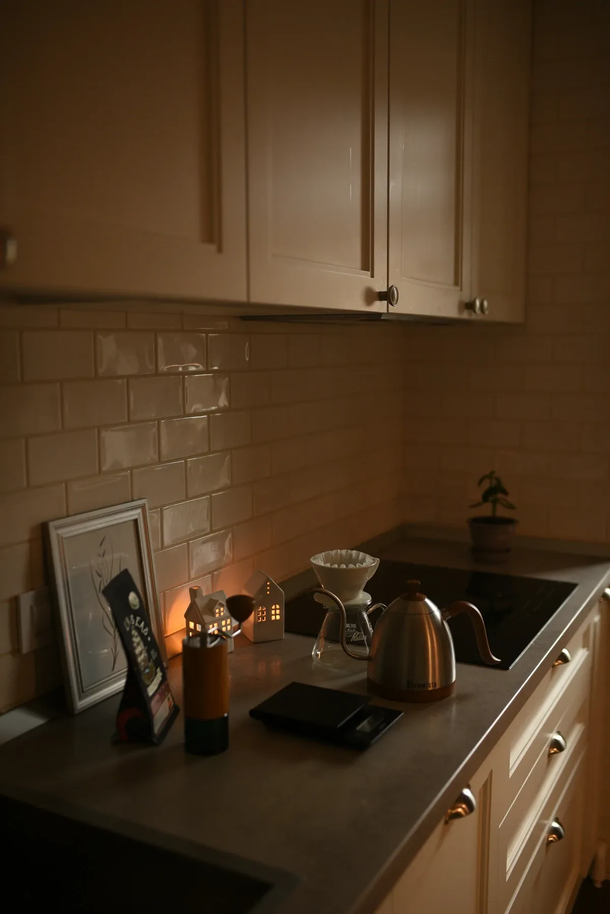 Dim kitchen corner with a lit candle, a framed print, a pour-over kettle and coffee gear on a dark counter under cream cabinets