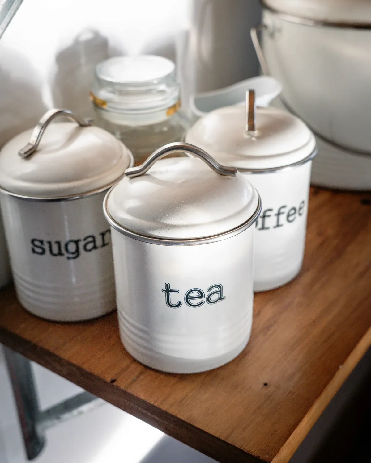 White enamel canisters labeled sugar, tea and coffee on a wooden wall shelf