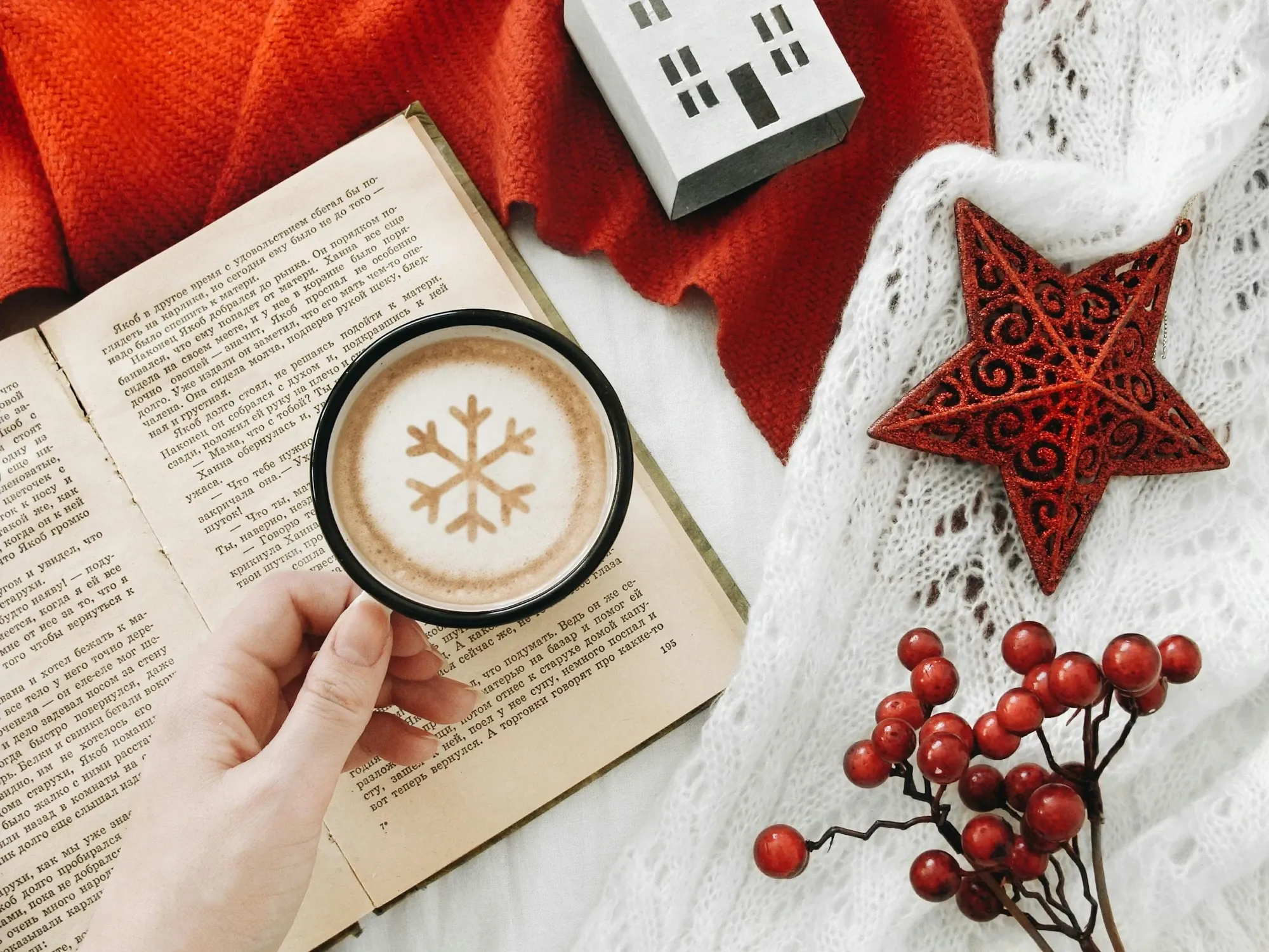 Hand holding a black mug of latte with a cocoa snowflake on the foam, over an open book with a red knit sweater and a red star ornament