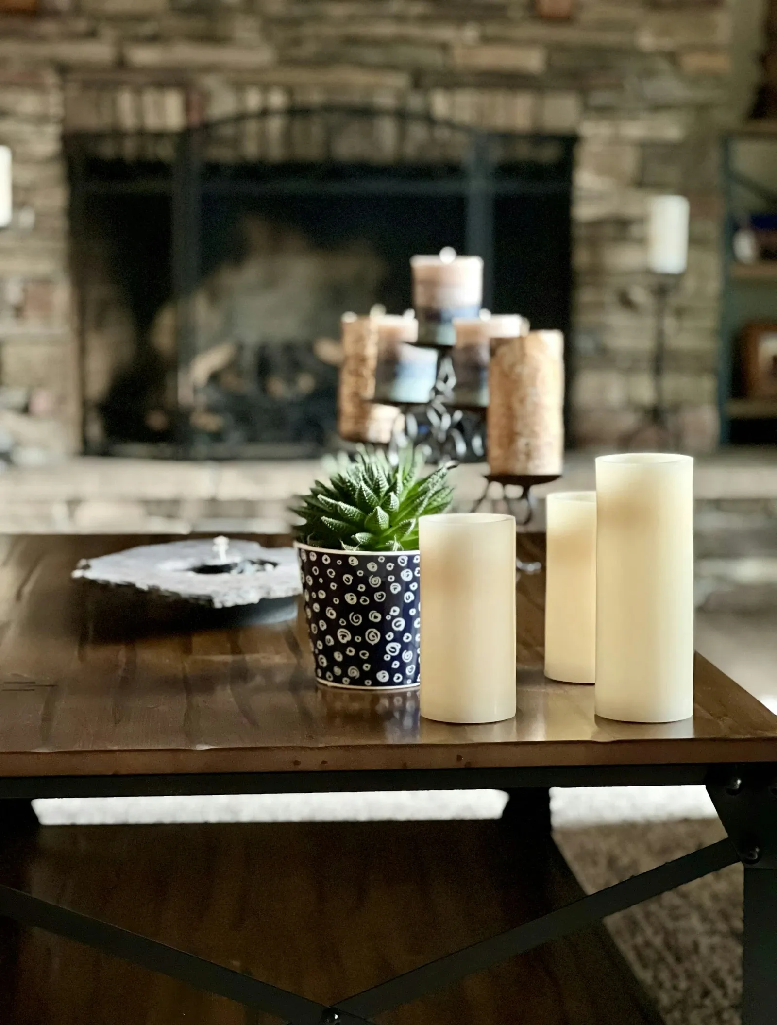 Cream flameless pillar candles and a succulent in a blue dotted pot on a dark wood table in front of a stone fireplace