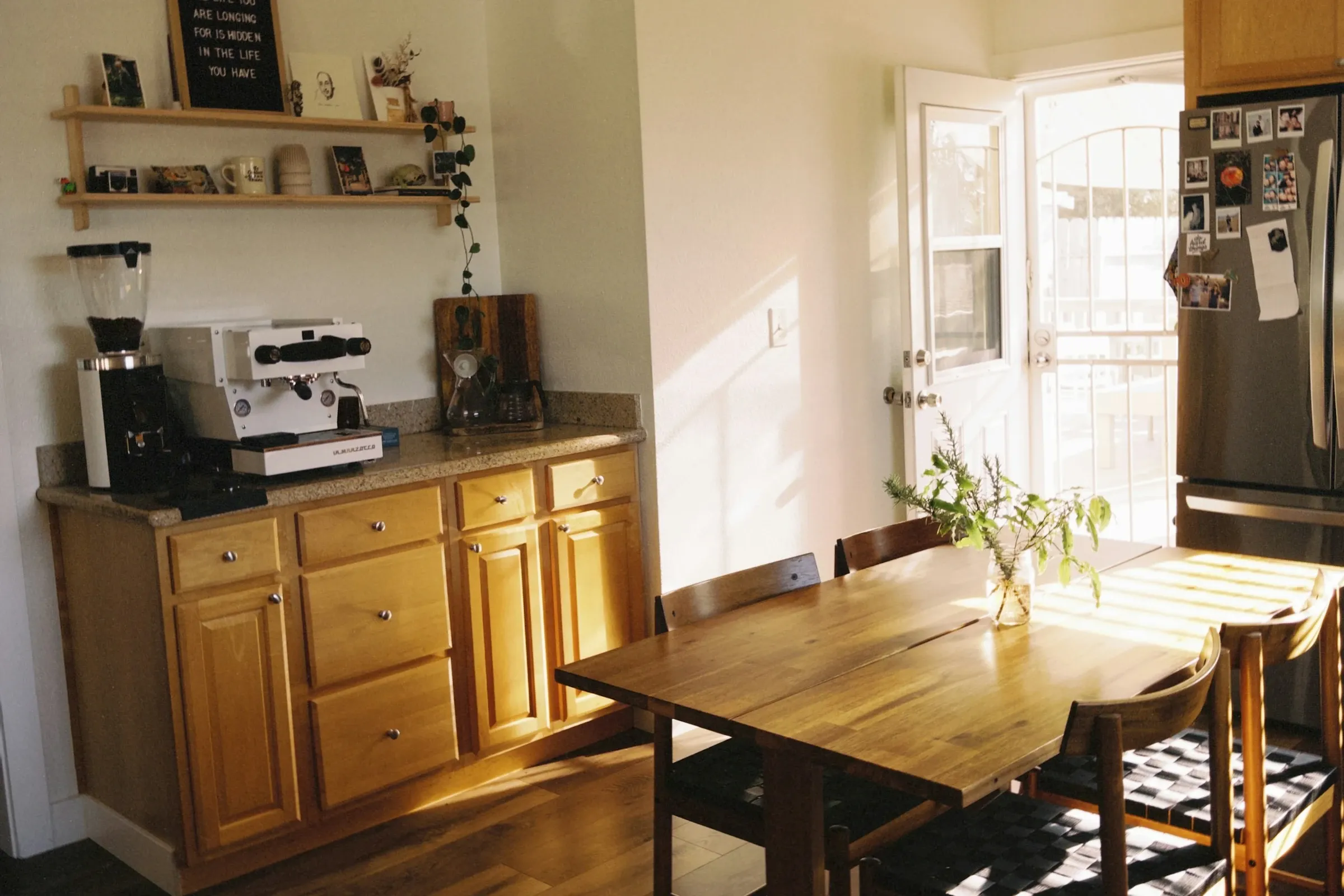 Sunlit kitchen with honey oak lower cabinets, an espresso machine in the corner, open wood shelves and a solid wood dining table