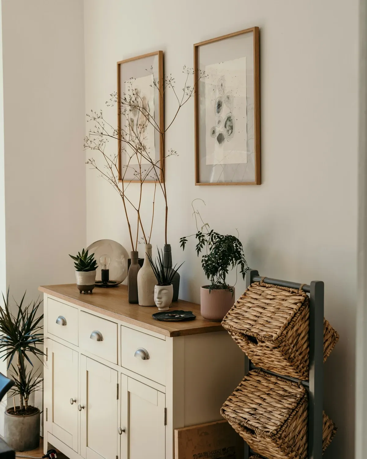 Light wood sideboard with wicker baskets on its open shelves, potted plants and vases on top against a white wall
