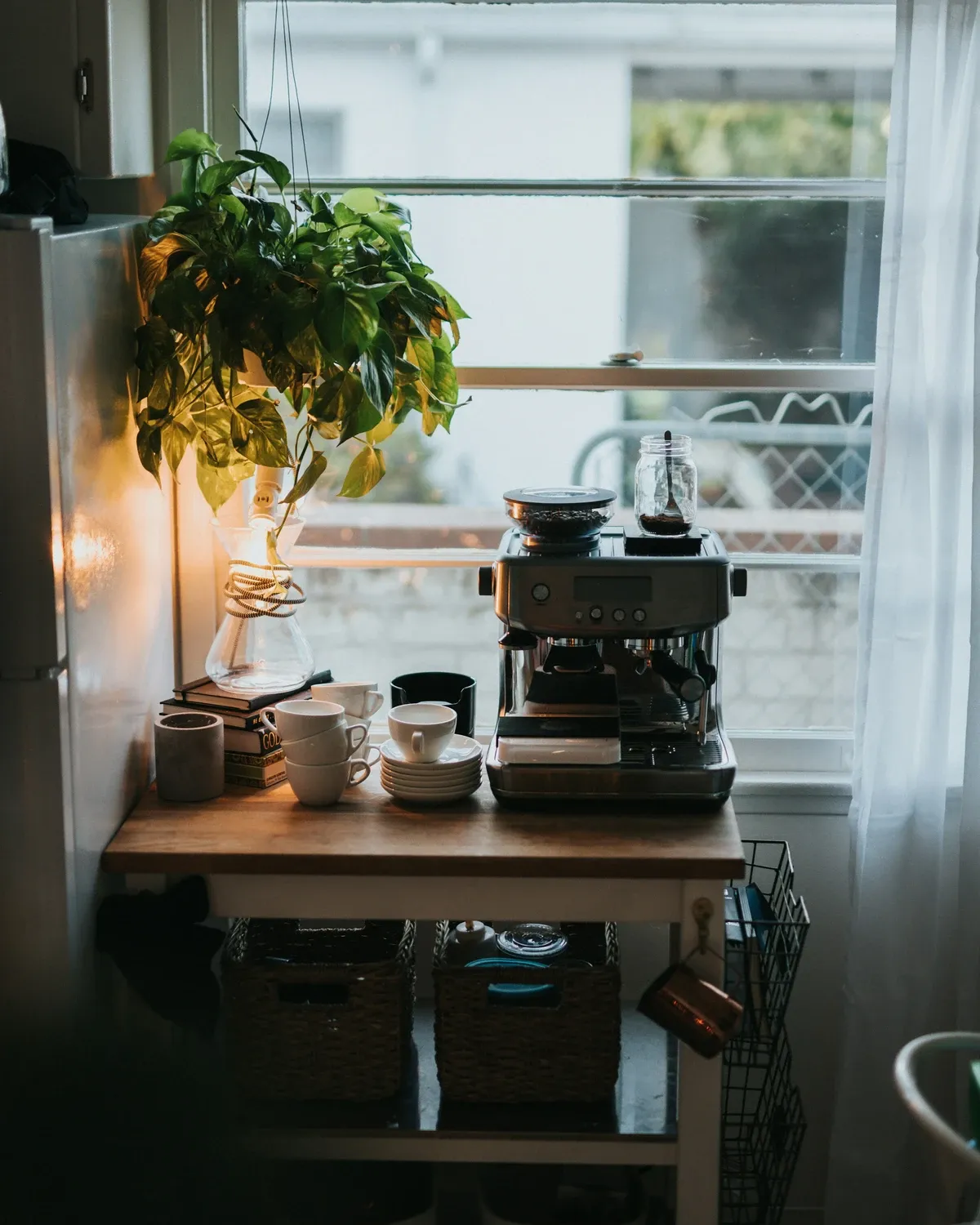 Small wood table by a bright window holding an espresso machine, a grinder and jars, with a plant on the sill