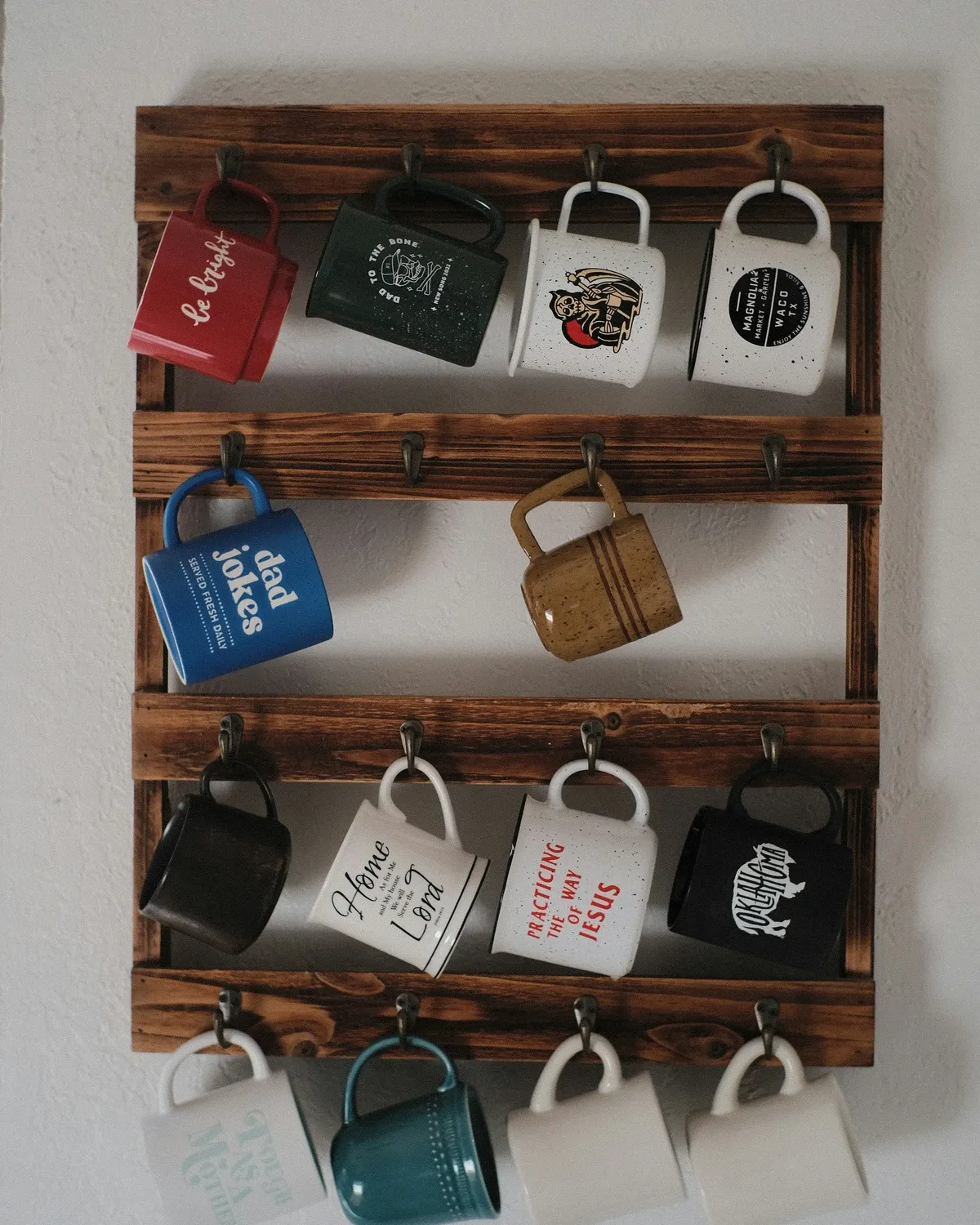 Dark stained wooden wall rack with three rows of hooks holding red, blue, black and white coffee mugs